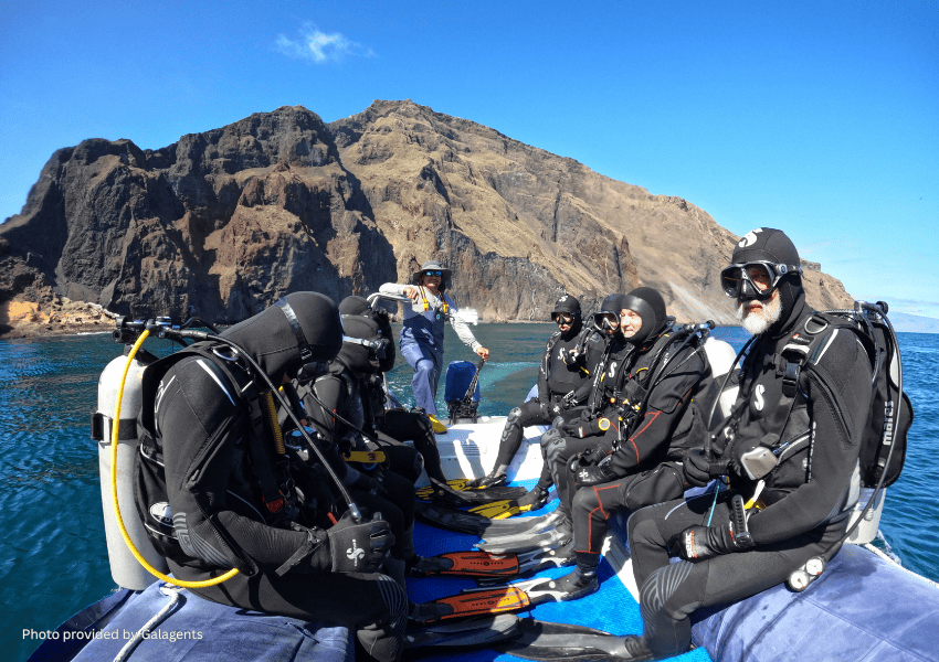 A group of scuba divers in full black wetsuits and gear sit on a small skiff, preparing for a dive near the rugged cliffs of the Galápagos Islands.