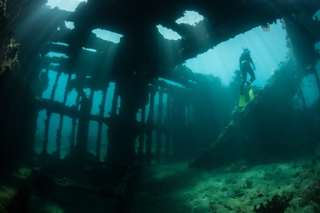 Diver exploring the interior of a sunken shipwreck with sunlight streaming through openings, creating a dramatic underwater scene