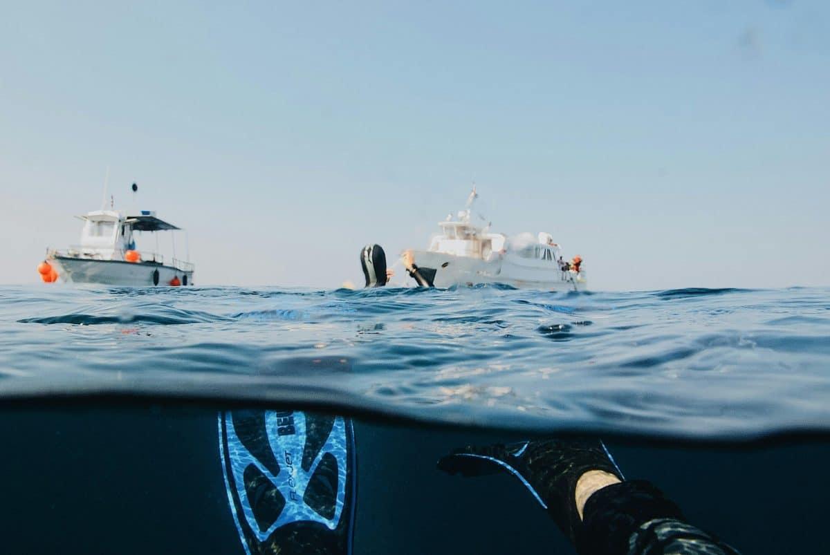 Split-level underwater photo showing a diver floating at the surface with fins clearly visible beneath, with dive boats anchored in the background