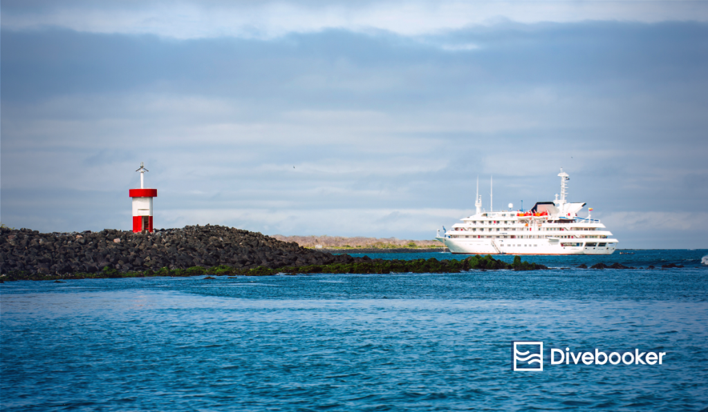 A large, white expedition cruise ship docked near a red and white lighthouse on a rocky outcrop in the Galápagos Islands, representing Galápagos diving tours.