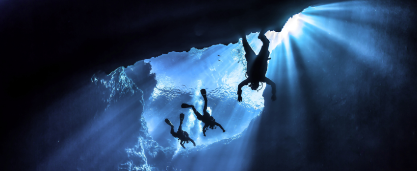 best-places-to-scuba-dive-advanced-cavern-experienc Three scuba divers silhouetted against dramatic light beams in a deep cavern, illustrating the challenging but rewarding environments found in the best places to scuba dive for advanced profile seekers.