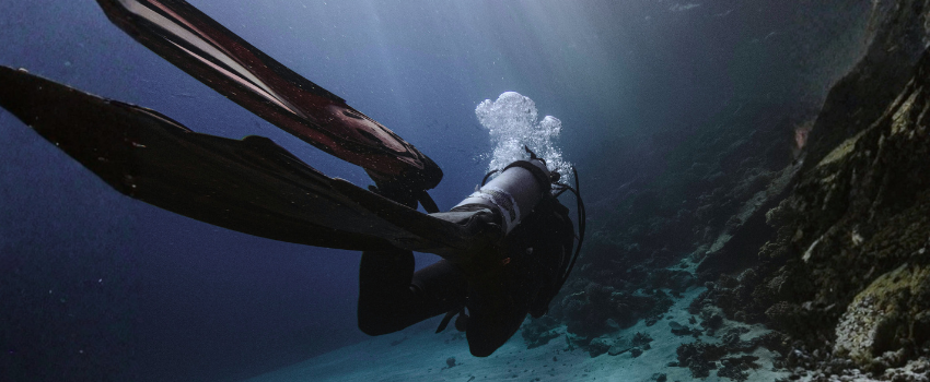 best-places-to-scuba-dive-specialist-gear-cold-water A scuba diver wearing full exposure protection and long fins swimming in deep blue water, representing the specialized gear strategy needed for the best places to scuba dive with varying temperatures.