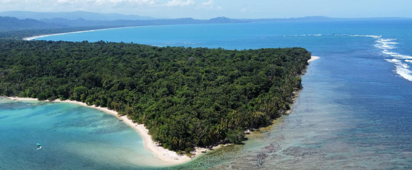 Aerial perspective of Cahuita National Park, showing a forested peninsula meeting white sandy beaches and a visible coral reef stretching into the Caribbean Sea.