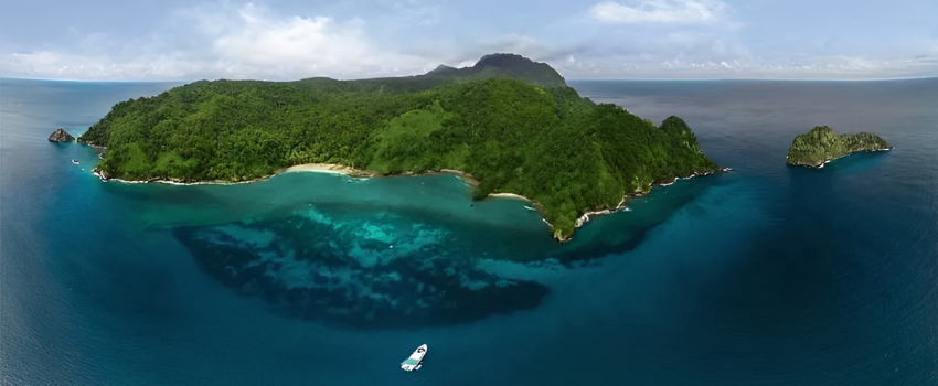 Panoramic aerial view of Cocos Island, one of the top dive destinations Costa Rica offers for hammerhead encounters. Lush green tropical rainforests surrounded by deep blue and turquoise ocean water with a single white boat anchored in the bay. 