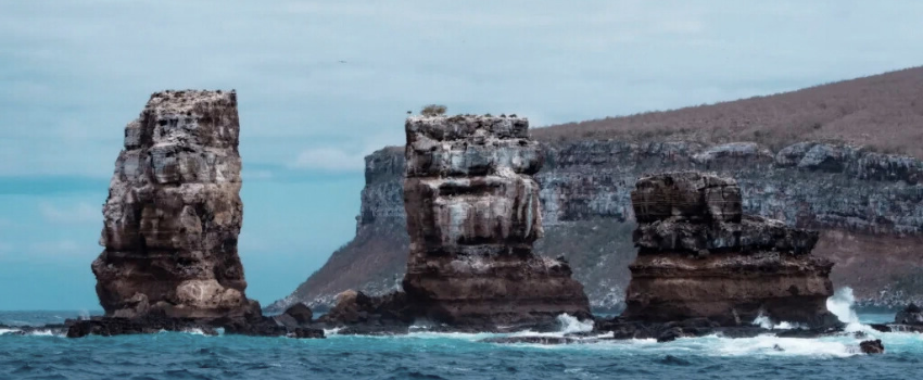 A coastal landscape featuring three large, rugged rock formations known as the Pillars of Evolution rising from the ocean, located in the Galápagos Islands