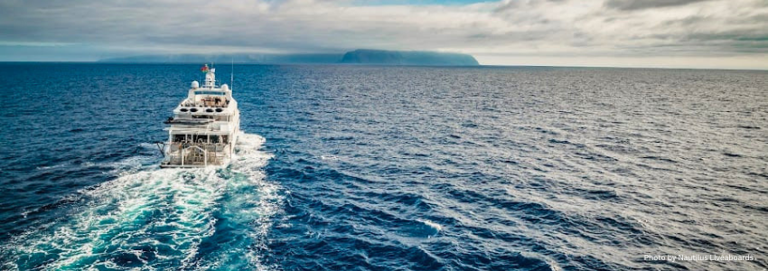 Nautilus luxury liveaboard vessel sailing near a rocky island, highlighting dive operators that offer flexible rebooking and transparent 2026 rates.