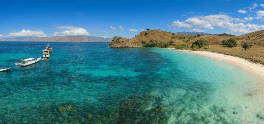 Pink Beach pano High-angle panoramic view of Pink Beach in Komodo, Indonesia, showcasing vibrant, crystal-clear turquoise ocean waters that reveal shallow coral reefs below.
